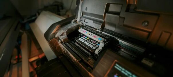 Zbliżenie na futurystyczną drukarkę przemysłową lub maszynę sterującą z odsłoniętymi wałkami, przyciskami, wyświetlaczem i ciepłym, przyciemnionym oświetleniem tworzącym techniczny nastrój.